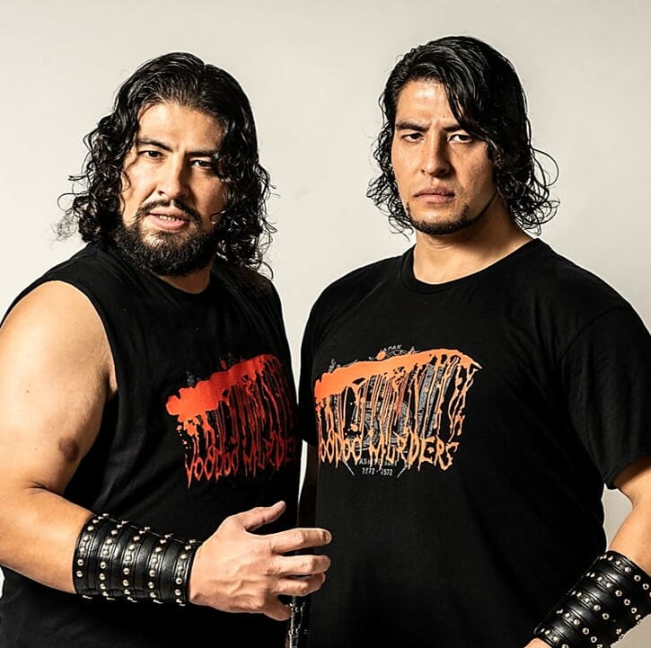 Superluchas - Dos luchadores con camisetas negras, GLEAT, parados uno al lado del otro durante la Nueva defensa de Saito Brothers.