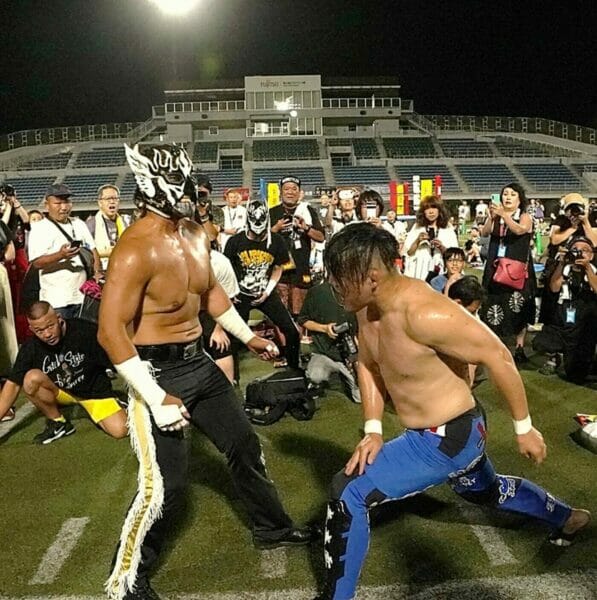 Dos luchadores (El Desperado y Hikaru Sato) se doblegan (derrotan) entre sí en un campo frente a una multitud.