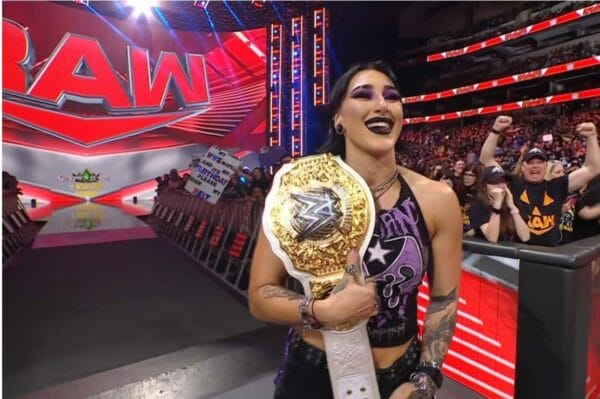 Superluchas - Drew McIntyre, una mujer, sostiene con confianza un cinturón en bruto frente a una multitud.