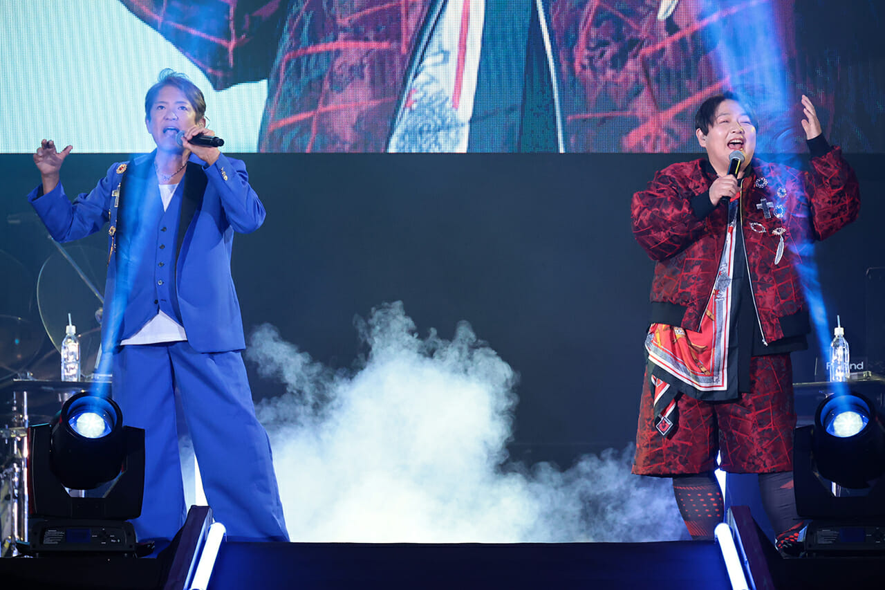 Superluchas - Dos hombres cantando en el escenario con humo saliendo de sus bocas durante una actuación de joshi.