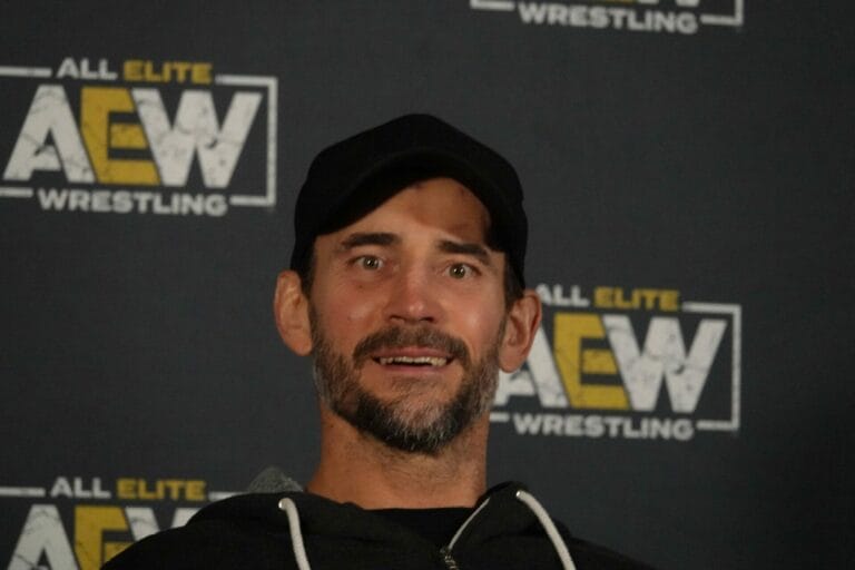 Superluchas - Un hombre con un sombrero negro está parado frente a un cartel de lucha libre de élite. ¿CM Punk en NJPW?