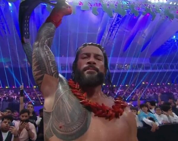 Superluchas - Roman Reigns, un luchador, sostiene una espada triunfalmente frente a una multitud que lo vitorea en Crown Jewel.