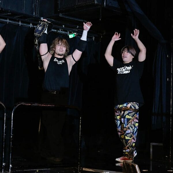 Superluchas - Tres luchadores, DDT, de pie en un escenario con las manos en alto.