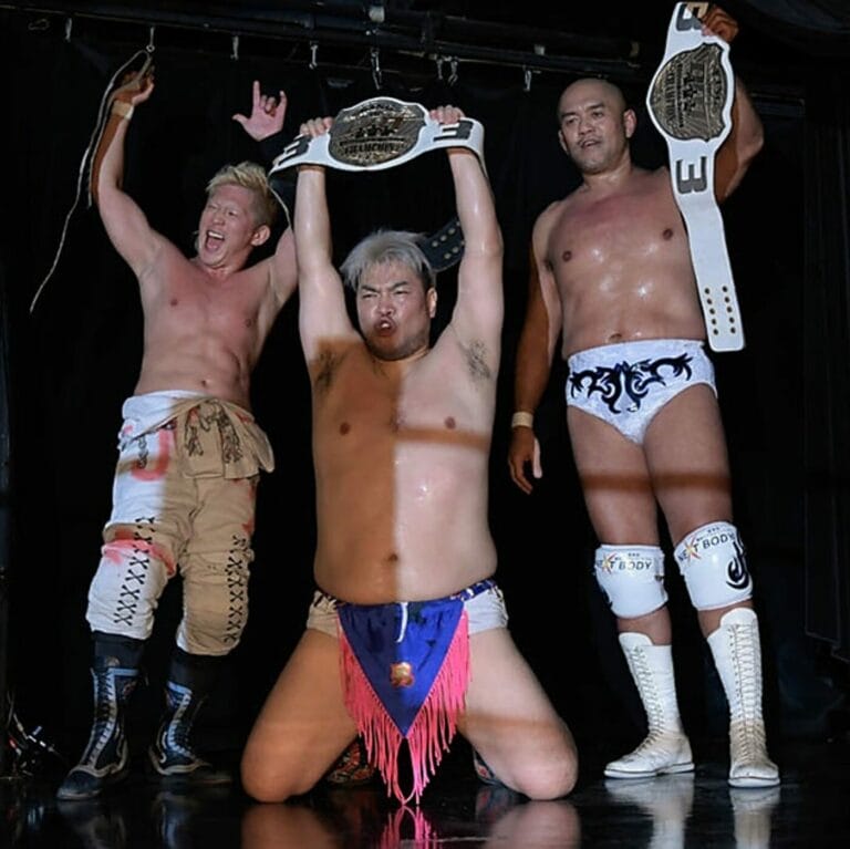 Superluchas - Tres luchadores que compiten en el torneo DDT, mostrando con orgullo sus cinturones en el escenario.