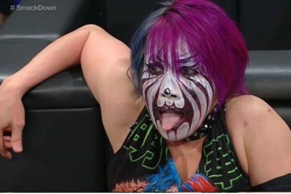 Superlucho - A woman with purple hair and makeup, Asuka, sitting on a sofa.