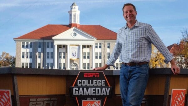 Superluchas - Kevin Dunn parado frente a un cartel del día del juego universitario en la WWE.