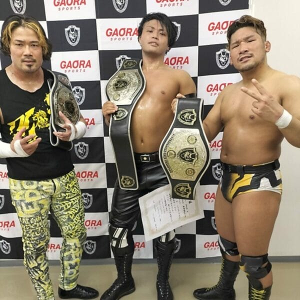 Superluchas - Tres luchadores de Z-Bratz, incluido Shun Skywalker, posan orgullosos con sus cinturones frente a una pared con la marca Dragon Gate.