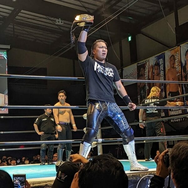 Superluchas - Un luchador, Daimonji So, sostiene triunfalmente el trofeo del Campeonato All Asia en un ring de lucha libre.