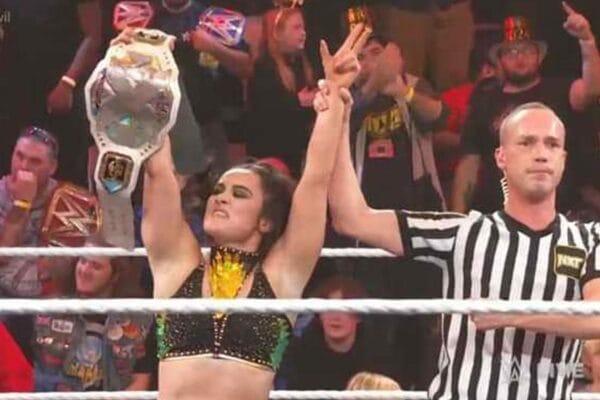 Superluchas - Lyra Valkyria, una mujer, sostiene triunfalmente un Campeonato de la WWE frente a un árbitro.